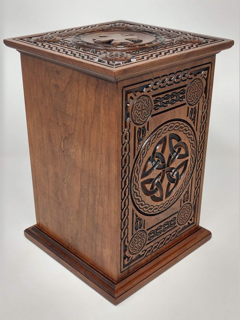 May include: A wooden box with intricate Celtic knotwork carvings. The box is made of dark wood and has a square shape. The top of the box features a Celtic knot design in the center.