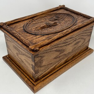 May include: A wooden box with a carved design on the lid. The box is made of dark wood with a natural finish. The lid is hinged and has a raised oval with a carved design of a fish.