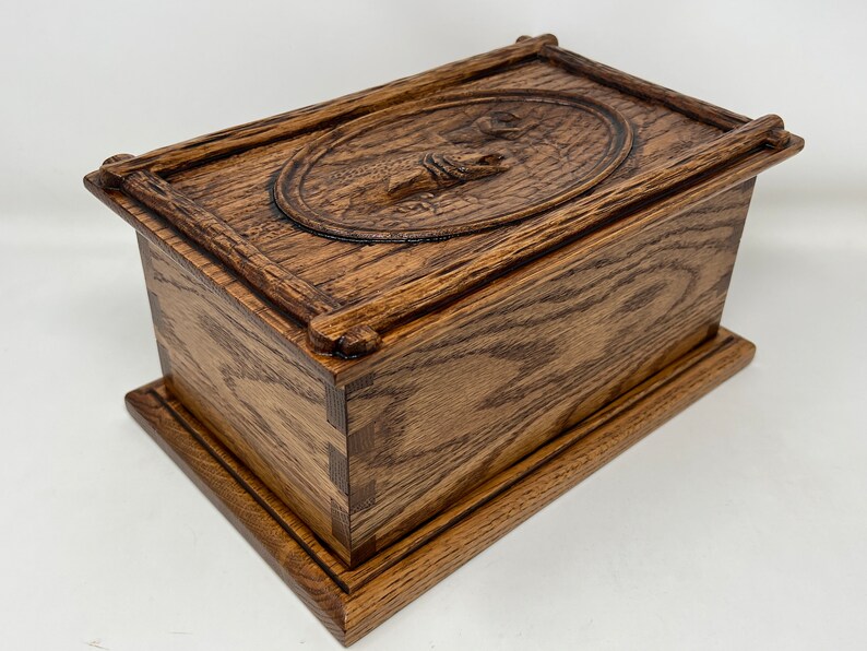 May include: A wooden box with a carved design on the lid. The box is made of dark wood with a natural finish. The lid is hinged and has a raised oval with a carved design of a fish.