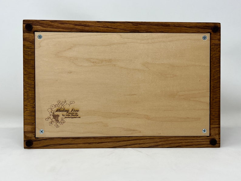 May include: A wooden box with a light wood interior and a dark wood frame. The box has a small engraved design on the inside that says "Abiding Love by John Shoba".