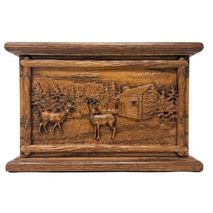 May include: A wooden box with a carved relief depicting a forest scene with two deer and a cabin. The box is rectangular with a dark brown finish and a decorative border. The overall theme is rustic and nature-inspired.