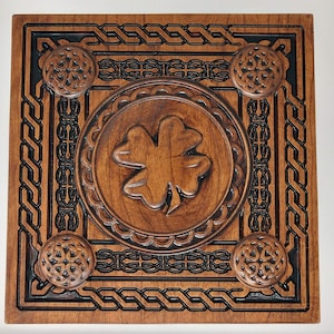 May include: A wooden square plaque with a Celtic knot border and a carved shamrock in the center. The shamrock is surrounded by a decorative circle.