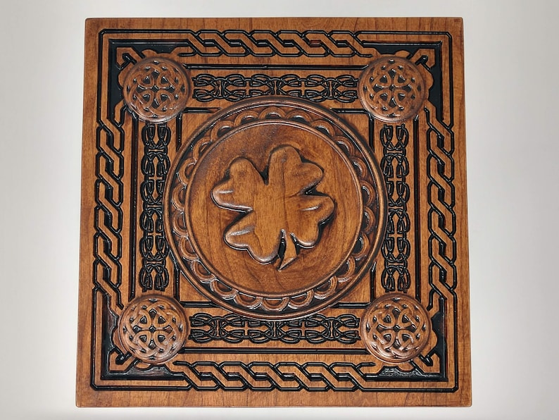 May include: A wooden square plaque with a Celtic knot border and a carved shamrock in the center. The shamrock is surrounded by a decorative circle.
