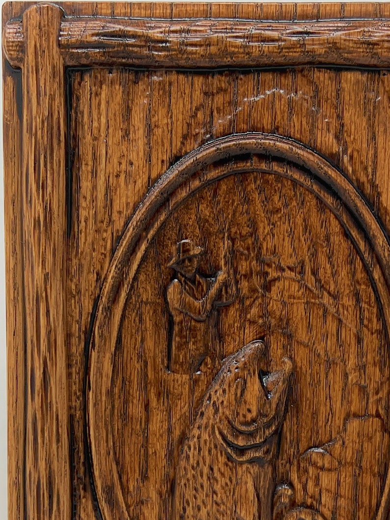 May include: A wooden plaque with a carved image of a fisherman holding a fishing rod and a large fish. The plaque is dark brown with a natural wood grain.