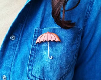 May include: A wooden brooch in the shape of an umbrella. The umbrella is red and has a silver handle. The brooch is pinned to a blue denim shirt.