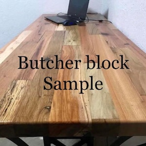 May include: Close-up of a wooden butcher block countertop. The wood has a natural, rustic look with variations in color and grain. The text "Butcher block Sample" is printed on the surface.