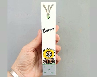 Pode incluir: Um marcador de livros branco com uma ilustração de leão pintada à mão. O leão tem uma juba amarela e está sentado na grama verde. O nome "Brenner" está escrito em letras pretas acima do leão.