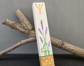 Pode incluir: Um item de madeira retangular pintado à mão com raminhos de lavanda. O design inclui flores roxas, caules verdes e um detalhe amarelo no topo. A seção inferior tem um padrão pontilhado marrom claro. Um pedaço de madeira flutuante repousa atrás do item.