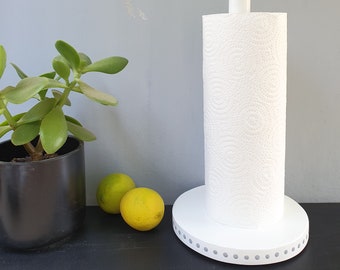 May include: White paper towel holder with a white base and a white rod. The base has a dotted pattern around the edge. A roll of white paper towels is on the rod. Two yellow lemons are on a black surface in the foreground.