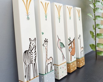 Pode incluir: Cinco blocos de madeira com ilustrações de animais pintadas. Cada bloco apresenta um animal diferente, incluindo uma zebra, uma girafa, um beija-flor, um bicho-preguiça e outra girafa. Os blocos são pintados em um fundo branco com detalhes verdes, marrons e amarelos.