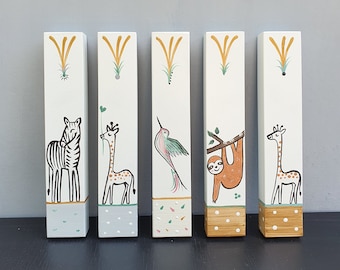 Pode incluir: Cinco blocos de madeira com ilustrações de animais pintadas. Os blocos apresentam uma zebra, uma girafa, um beija-flor, um preguiça e outra girafa. Cada bloco tem uma base de cor diferente e um desenho amarelo na parte superior.