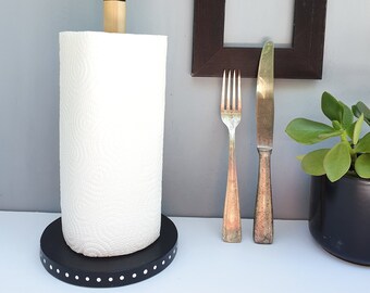 May include: A black and white paper towel holder with a wooden dowel and a white paper towel roll. The holder has a black base with white polka dots. A black picture frame, a fork, and a knife are on the wall behind the holder.