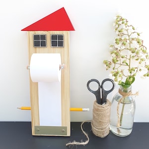 May include: A wooden house-shaped memo board with a red roof, two black windows, and a roll of paper attached to the front. A pencil is placed on the board, and a spool of twine is in the foreground.
