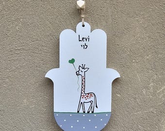 Pode incluir: Uma mão de Hamsa de madeira branca com um padrão de bolinhas azuis e brancas na parte inferior. A mão de Hamsa tem uma girafa com um balão verde e o nome "Levi" escrito em hebraico e inglês.