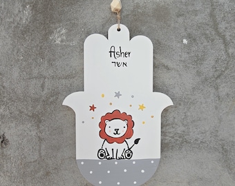 Pode incluir: Uma decoração de parede em forma de mão Hamsa de madeira branca com o nome "Asher" em inglês e hebraico. Apresenta uma ilustração de leão de desenho animado com uma juba vermelha, sentado em uma base cinza pontilhada e é adornado com estrelas. Uma conta de madeira e uma corda são anexadas para pendurar.