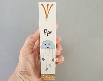 Pode incluir: Um marcador de página de madeira branco com uma nuvem azul sorridente e estrelas penduradas nela. O nome "Ron" está escrito no marcador de página.