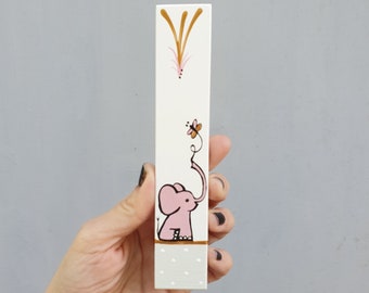 Pode incluir: Um bloco de madeira branco com um design de elefante rosa. O elefante tem uma tromba longa que chega a uma borboleta rosa. O fundo é branco com detalhes dourados.
