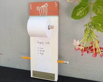 Pode incluir: Um quadro de avisos de madeira branco com um design de cacto vermelho e verde. O quadro tem um rolo de papel e um lápis presos a ele. O papel tem as palavras "Shopping List:" e "Love, Kisses, Hugs" escritas nele.