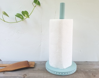May include: A light blue paper towel holder with white polka dots. The holder has a wooden base and a metal rod to hold the paper towel roll. The holder is sitting on a wooden surface.