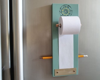 Pode incluir: Um quadro magnético azul-verde com um suporte para rolo de papel e um lápis. O quadro tem um design decorativo com um padrão de mandala e uma placa de metal dourado.