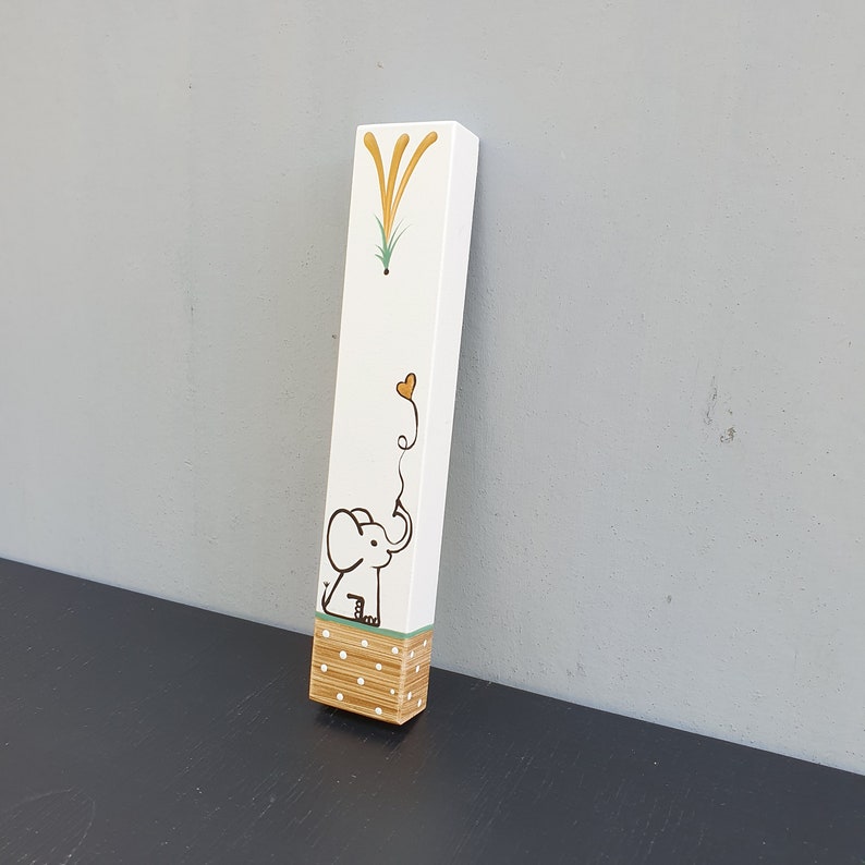 May include: A white wooden block with a hand-painted design of an elephant holding a heart-shaped balloon. The elephant is black and the balloon is yellow. The block has a green and white polka dot pattern at the bottom.
