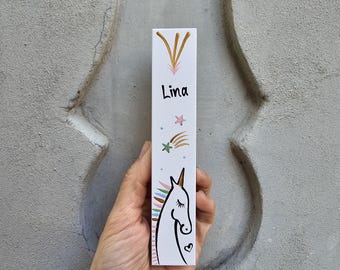 Pode incluir: Bloco de madeira branco com uma ilustração de unicórnio, uma estrela dourada e o nome "Lina" escrito em preto. O unicórnio tem uma crina e uma cauda arco-íris.