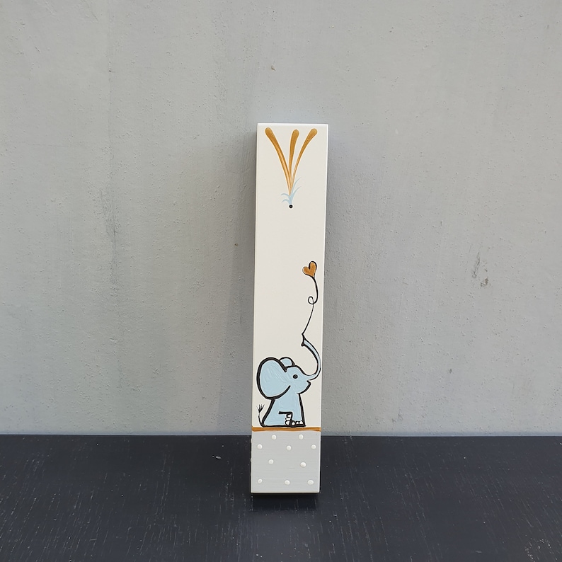 May include: A white wooden sign with a blue elephant holding a heart-shaped balloon. The elephant is painted in a whimsical style with a brown outline. The sign has a white polka dot pattern.