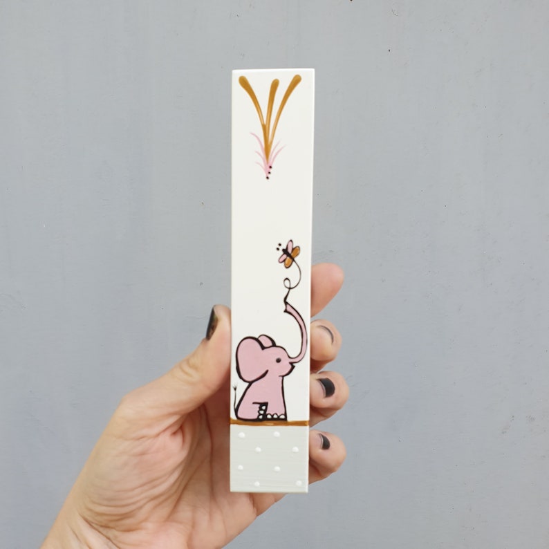 May include: A white wooden block with a pink elephant design. The elephant has a long trunk that is curled up and reaching towards a pink butterfly. The block has a gold and white polka dot pattern at the bottom.
