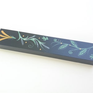 May include: A black rectangular box with a painted design of green leaves and gold accents. The design features a stylized branch with leaves and swirls.