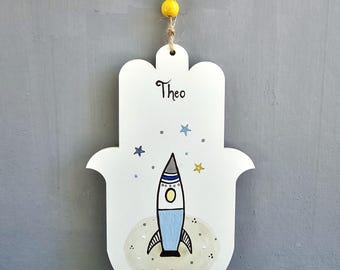 Pode incluir: Enfeite de parede em forma de mão de Hamsa em madeira branca com um desenho de foguete pintado. O foguete é azul, cinza e branco, com estrelas amarelas. O nome "Theo" está escrito em itálico preto.