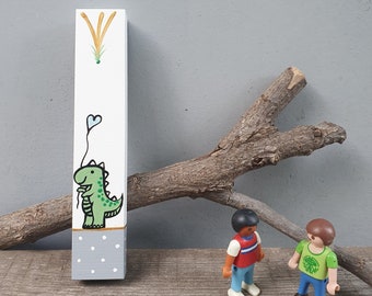Pode incluir: Um bloco de madeira pintado de branco com um dinossauro verde segurando um balão em forma de coração azul. O bloco tem um padrão de bolinhas cinza e brancas na parte inferior. O bloco está sentado em uma superfície de madeira.