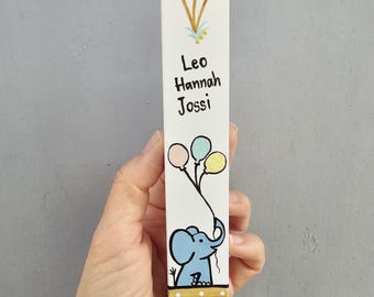 Pode incluir: Marcador de livros de madeira pintado à mão com um elefante azul segurando três balões. O marcador de livros tem os nomes "Leo Hannah Jossi" e detalhes dourados e azuis. A parte inferior tem um design de bolinhas douradas e brancas.