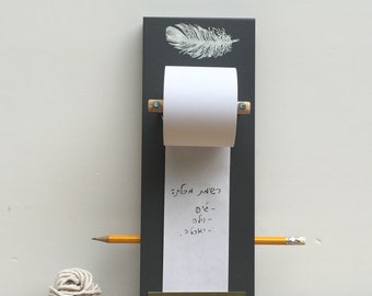 Pode incluir: Um quadro de avisos de madeira cinza com um design de pena branca. O quadro tem um rolo de papel com um porta-lápis e uma placa de latão na parte inferior. O papel tem notas escritas à mão em hebraico.