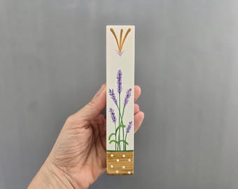 Pode incluir: Um bloco de madeira retangular pintado à mão com um desenho de lavanda. O bloco tem um fundo creme com flores de lavanda roxas e caules verdes. A seção inferior é marrom com bolinhas brancas.