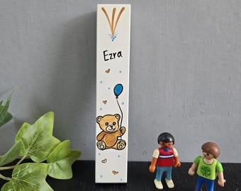 Pode incluir: Uma régua de crescimento retangular de madeira branca com um urso de pelúcia pintado à mão segurando um balão azul. O nome "Ezra" está escrito em letra preta. Pequenos corações castanhos e pontos azuis decoram a régua. Artigo decorativo.