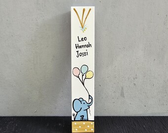 Pode incluir: Um letreiro de madeira branco com parte superior e inferior douradas. O letreiro tem um elefante pintado à mão segurando balões nas cores rosa, azul e amarelo. O letreiro também tem os nomes Leo, Hannah e Jossi escritos em preto.