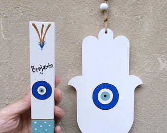 Pode incluir: Mão de Hamsa de madeira branca e objeto retangular com um design de olho grego. O objeto retangular tem o nome "Benjamin" escrito. Ambos os itens são pintados com detalhes em azul e castanho.