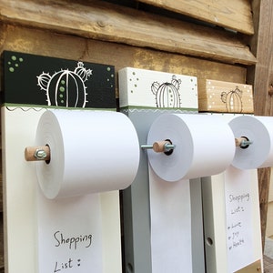 May include: Three wooden memo boards with a cactus design. Each board has a roll of white paper attached to it. The paper is labeled "Shopping List" and has handwritten items on it. The boards are painted in black, white, and brown.