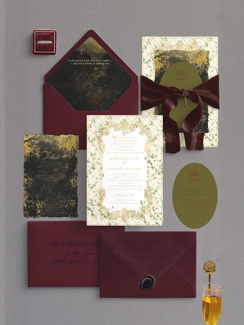 May include: A collection of wedding stationery in burgundy, gold, and olive green. Includes invitations, envelopes, and a ring box. The invitation features the names "Gabrielle Noel" and "Gilbert Soriano". A glass of champagne with a gold rose stirrer is also present.