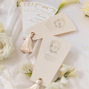 May include: Ivory wedding program fans with gold foil accents and tassel details. The fans feature the names "Macey & David" and the wedding date, January 26, 2026. The image includes white flowers and a soft, white fabric background.