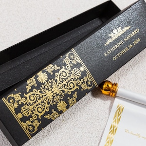 Puede incluir: Una caja negra con detalles decorativos dorados y el texto "KATHERINE NAVARRO OCTOBER 26, 2024". La caja contiene un bolígrafo blanco con punta dorada. La caja tiene un patrón dorado decorativo.