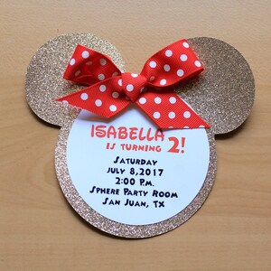 May include: A Mickey Mouse-themed birthday invitation. The invitation is shaped like Mickey's head, with gold glitter ears and a red bow with white polka dots. The invitation reads "Isabella is turning 2!" with party details.