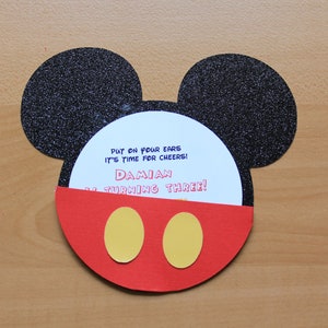 May include: A Mickey Mouse-themed birthday invitation. The invitation is shaped like Mickey's head, with black glitter ears, a red face, and yellow circles. The invitation reads: "Put on your ears, it's time for cheers! Damian is turning three!"