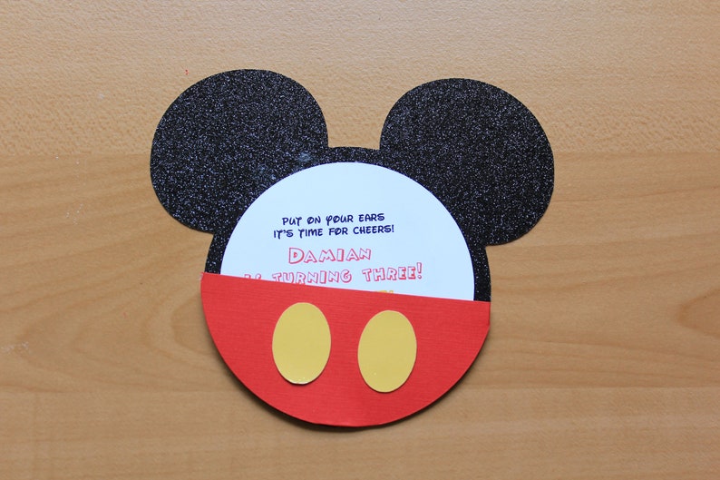 May include: A Mickey Mouse-themed birthday invitation. The invitation is shaped like Mickey's head, with black glitter ears, a red face, and yellow circles. The invitation reads: "Put on your ears, it's time for cheers! Damian is turning three!"