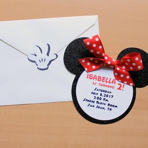 May include: A white envelope with a blue Mickey Mouse hand print and a Minnie Mouse-themed invitation. The invitation is shaped like Minnie's head, with black glitter ears and a red bow with white polka dots. The invitation reads "Isabella is turning 2!" with the date and location.
