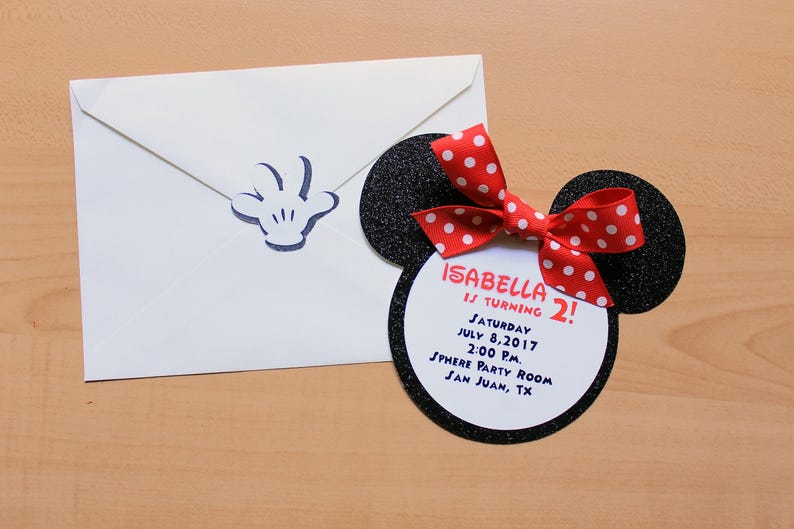 May include: A white envelope with a blue Mickey Mouse hand print and a Minnie Mouse-themed invitation. The invitation is shaped like Minnie's head, with black glitter ears and a red bow with white polka dots. The invitation reads "Isabella is turning 2!" with the date and location.