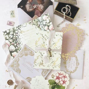 Può includere: Un assortimento di partecipazioni di nozze, tra cui inviti, biglietti di risposta e buste. Gli inviti presentano decorazioni floreali e dorate, con un dipinto vintage sulla busta. È presente anche una scatola decorativa con una collana di perle e una chiave legata con un nastro.