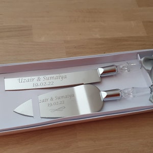 May include: A silver cake knife and server set in a white box with two silver bows. The knife and server are engraved with the names "Uzair & Sumaiya" and the date "19.02.22".