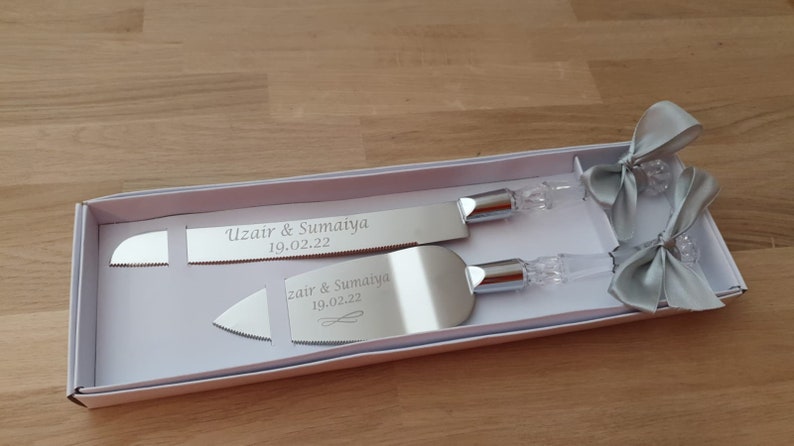 May include: A silver cake knife and server set in a white box with two silver bows. The knife and server are engraved with the names "Uzair & Sumaiya" and the date "19.02.22".