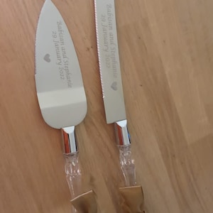 May include: A set of two silver cake knives with gold ribbon bows. The knives are engraved with the text "Babisan and Stephanie 29 January 2022".